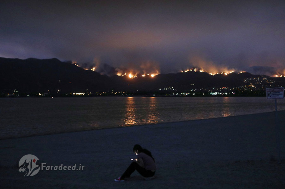 زنی در کنار رودخانه در حالی که جنگل‌های کالیفرنیا در آتش می‌سوزند، تلفن همراه خود را چک می‌کند